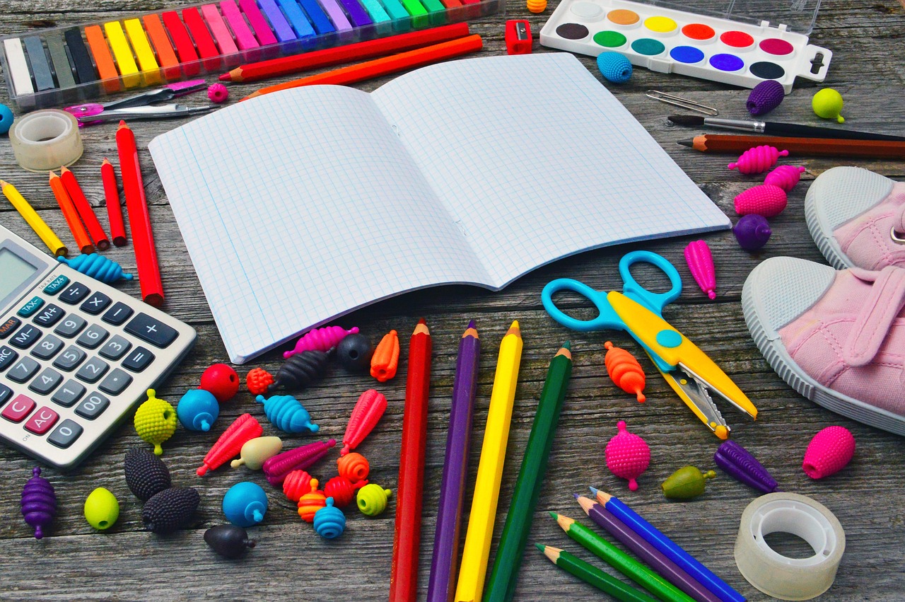 A flat lay on a weathered wooden surface displays an open blank grid-lined notebook surrounded by a vibrant assortment of school supplies: colorful pencils, crayons, a watercolor paint set, pink sneakers, a calculator, scissors, and various novelty erasers.